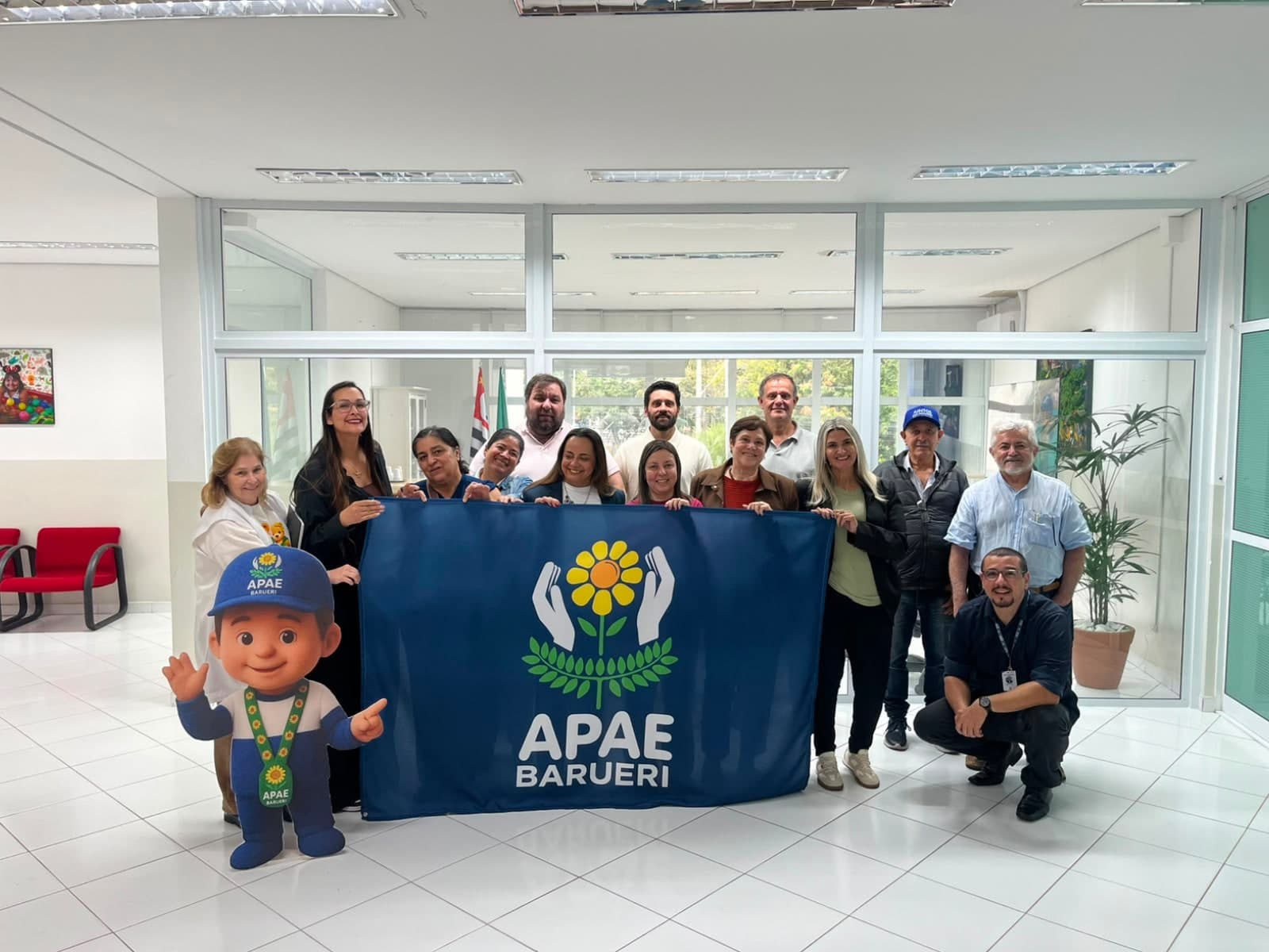 Diretoria, conselhos e equipe da APAE Barueri reunidos no hall da sede, segurando a bandeira da instituição, ao lado do mascote Apaezito.
