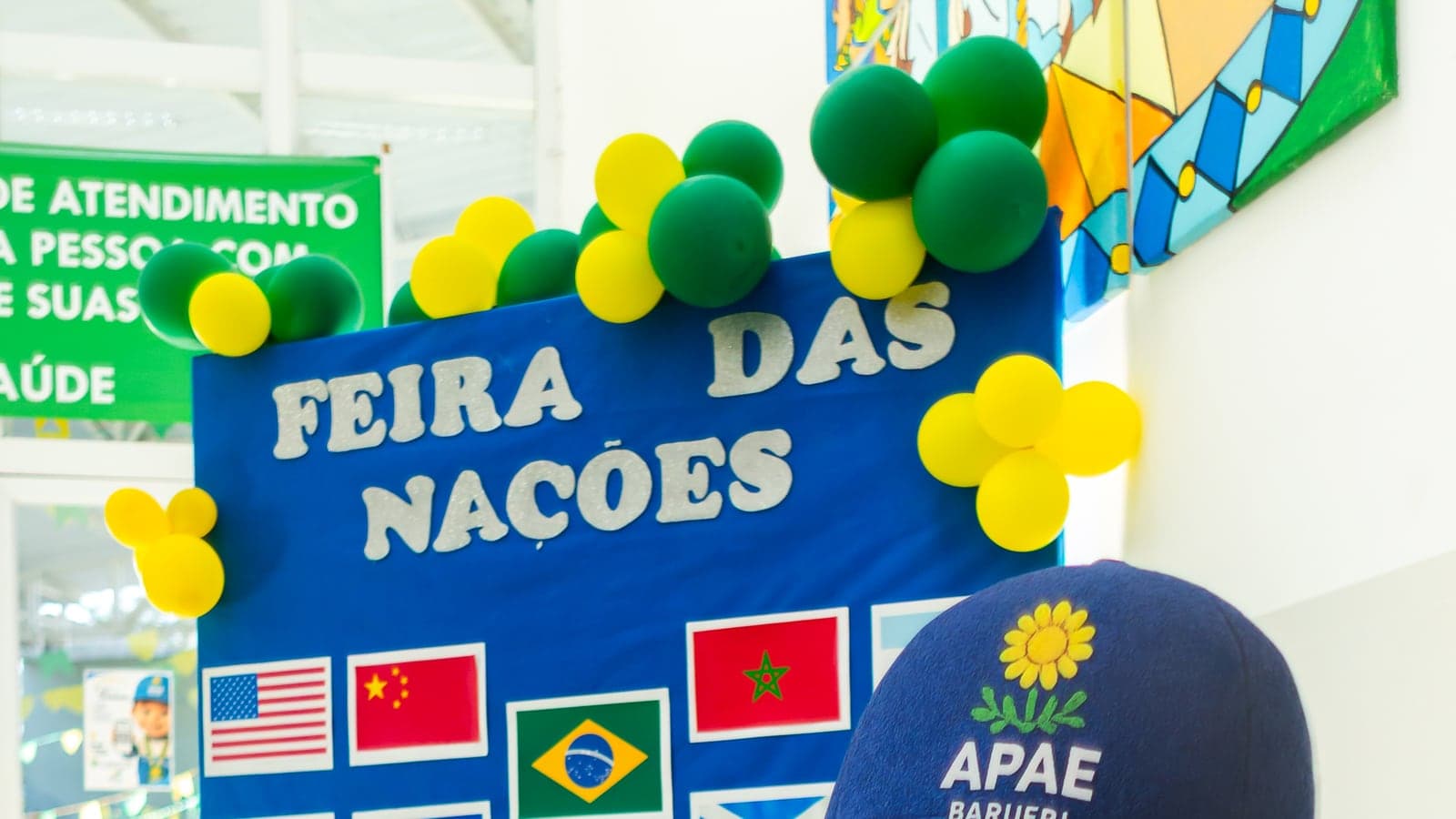 Painel da 1ª Feira das Nações da APAE Barueri com bandeiras de vários países, balões e o mascote Apaezito