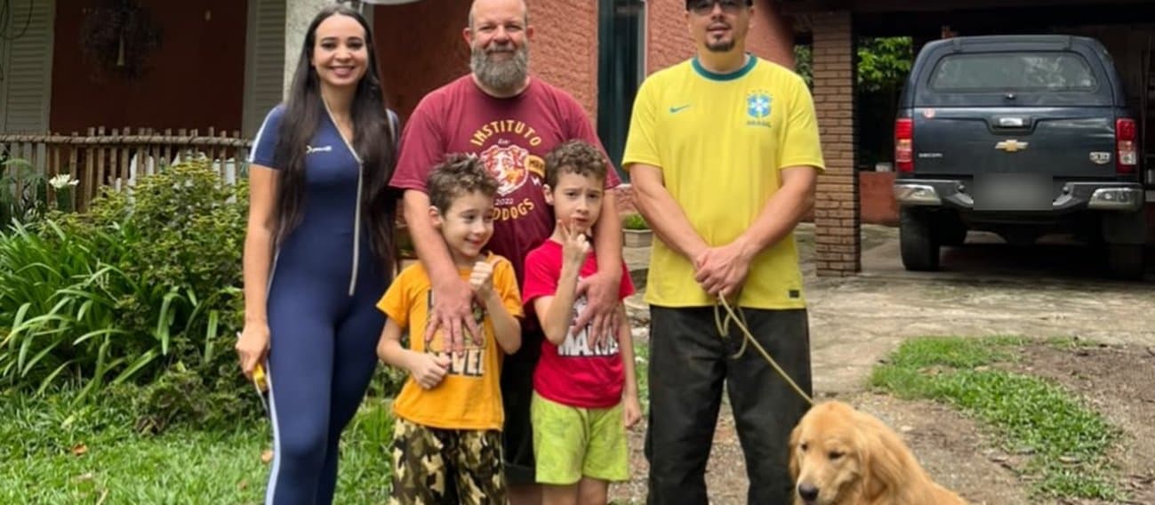 Diante de uma casa de tijolos aparentes cercada de plantas, sede do Instituto Red Dogs, um grupo posa para a foto: uma mulher de macacão azul, um homem de barba com camiseta do instituto, que apoia as mãos nos ombros de dois meninos, e um homem de boné e camisa amarela que segura pela guia um cão golden retriever, sentado na grama ao lado dele.