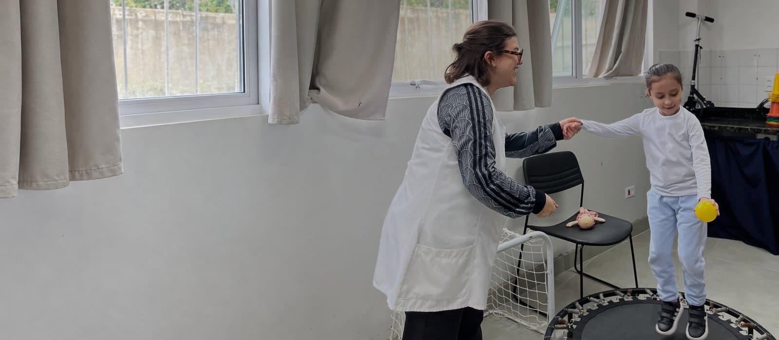 Numa sala de atendimento ampla, com janelas e cortinas bege, uma profissional de colete branco segura a mão de uma menina que pula num minitrampolim, com uma bolinha amarela na mão.