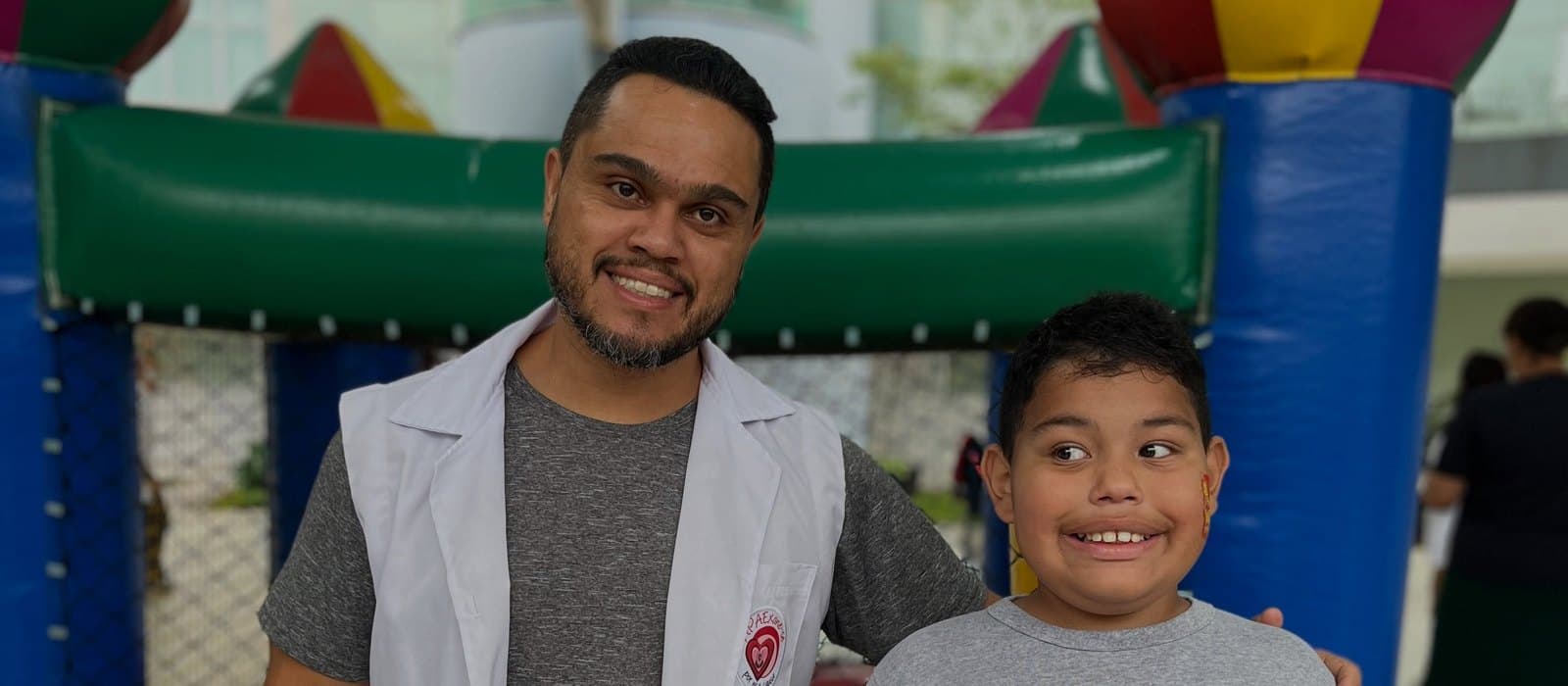 Um profissional da APAE Barueri, de jaleco branco, sorri ao lado de um menino atendido, com a mão em seu ombro, diante de um brinquedo inflável colorido no pátio da sede.
