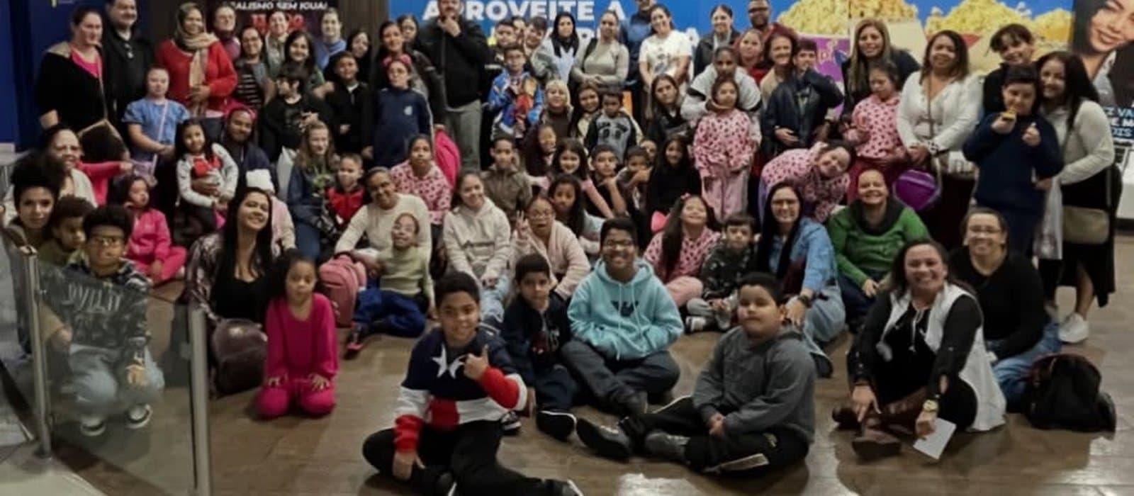 Grande grupo de crianças, adolescentes e adultos reunidos para uma foto no saguão de um cinema, alguns sentados no chão e outros de pé, diante de um painel de pipoca e da placa de uma sala Macro XE da Cinépolis.