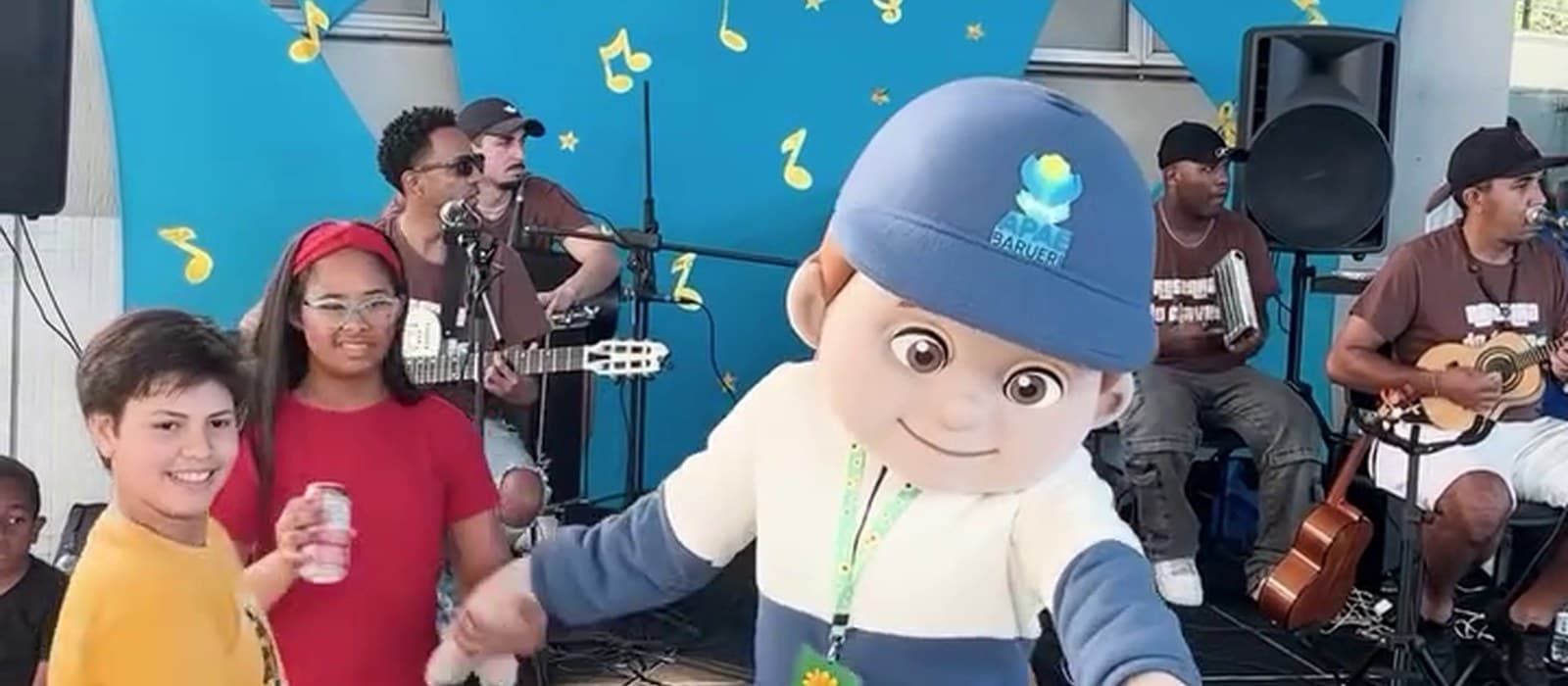 O Apaezito, mascote da APAE Barueri, dança de braços abertos rodeado por crianças, diante do palco onde o grupo toca; ao fundo, painel azul com balões azuis e amarelos.