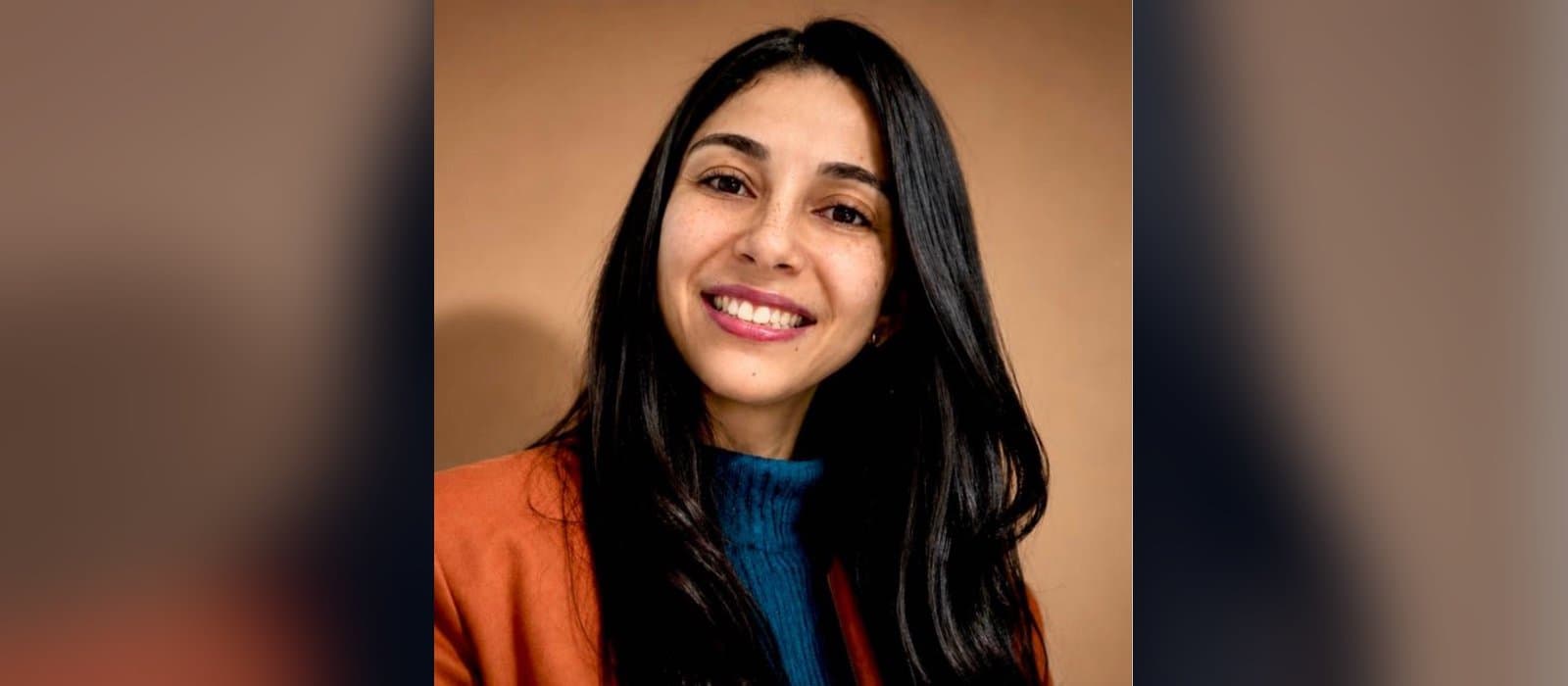 Retrato da psicóloga Henriette Russo Luiz de Barros, docente do curso: mulher de cabelos longos, pretos e lisos, sorrindo para a câmera, de blusa de gola alta azul-petróleo e casaco cor de ferrugem, diante de uma parede bege.