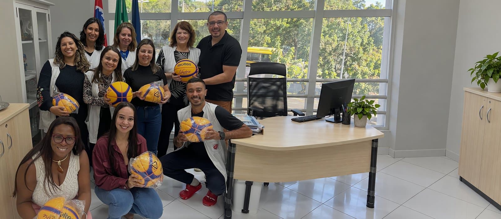Dez integrantes da equipe da APAE Barueri, a maioria de colete branco, sorriem numa sala com bandeiras ao fundo segurando bolas de basquete amarelas e azuis, várias ainda na embalagem plástica.