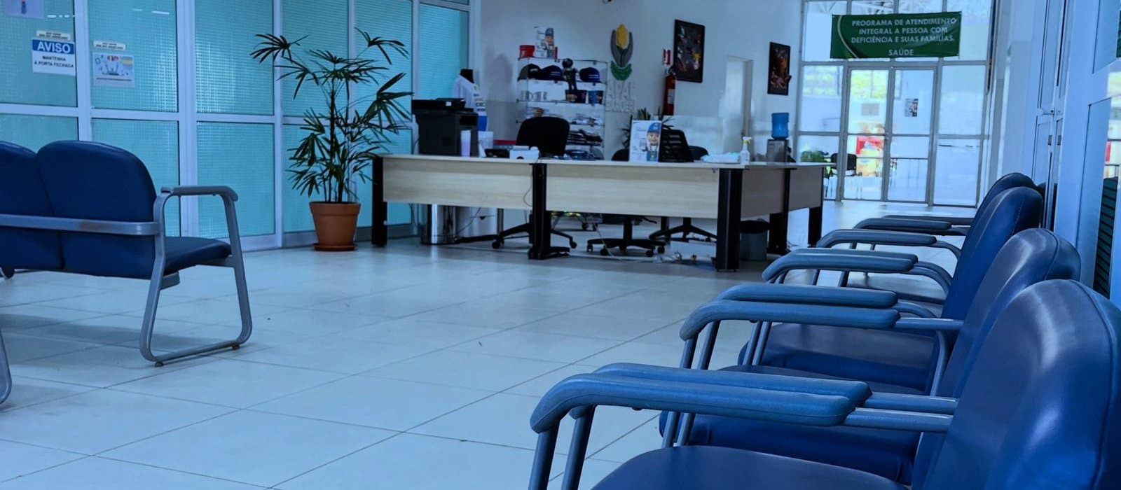 Salão de entrada da APAE Barueri com fileiras de cadeiras azuis, uma palmeira em vaso, o balcão de atendimento em madeira clara ao fundo e uma faixa verde com a inscrição “Programa de atendimento integral à pessoa com deficiência e suas famílias”.