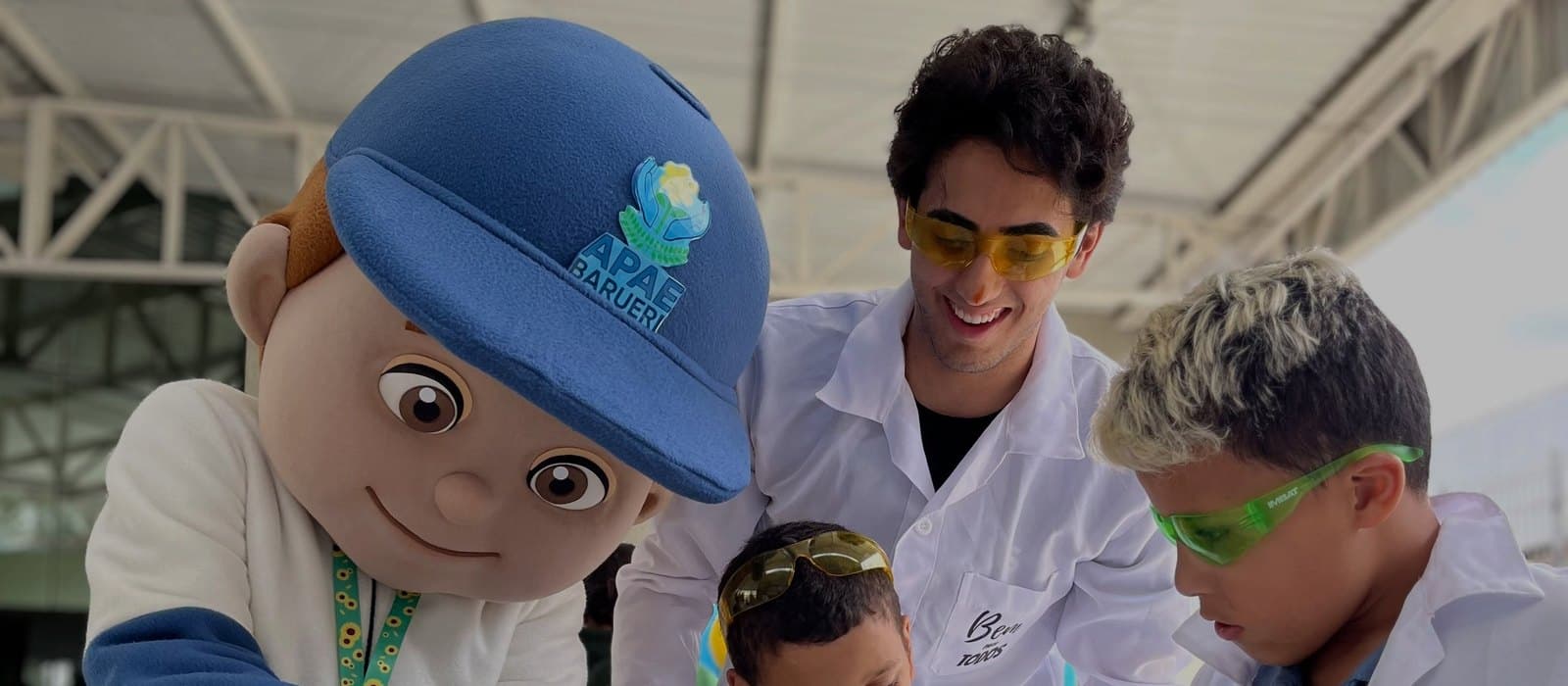 No pátio coberto da APAE Barueri, o APAEzito, personagem de boné azul da instituição, despeja um líquido de um pequeno frasco numa bandeja de metal com copinhos azuis. Ao lado, dois atendidos de jaleco branco acompanham a experiência de química, um deles com óculos de proteção verdes, e um voluntário de jaleco e óculos amarelos sorri atrás deles.