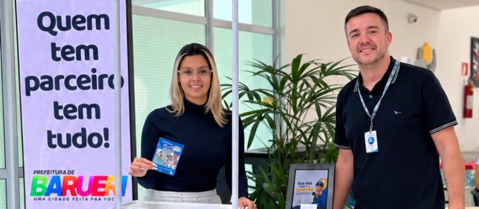 Dois colaboradores da APAE Barueri sorriem na recepção da sede ao lado dos novos equipamentos: à direita, o totem branco da Ouvidoria Geral, com tela no alto, a frase “Sua opinião é muito importante para nós!” e o símbolo da APAE Barueri; à esquerda, um telão vertical que exibe a mensagem “Quem tem parceiro tem tudo!”; ao centro, um balcão da campanha Nota Fiscal Paulista da APAE Barueri e uma palmeira em vaso.