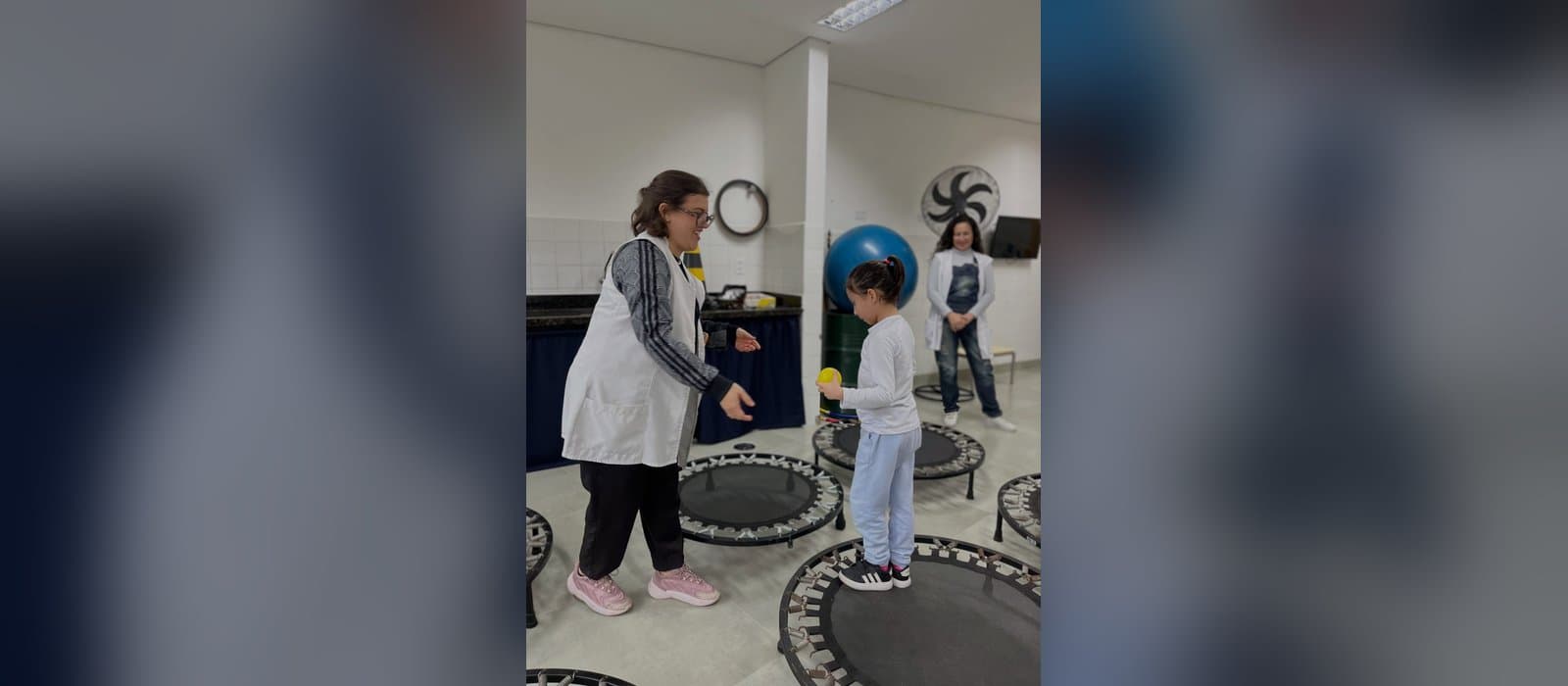 Numa sala de atendimento com vários minitrampolins redondos no chão, uma menina de blusa branca, de pé sobre um deles, segura uma bolinha amarela, enquanto uma profissional de avental branco estende as mãos para ela; ao fundo, uma bola suíça azul e outra profissional observando.
