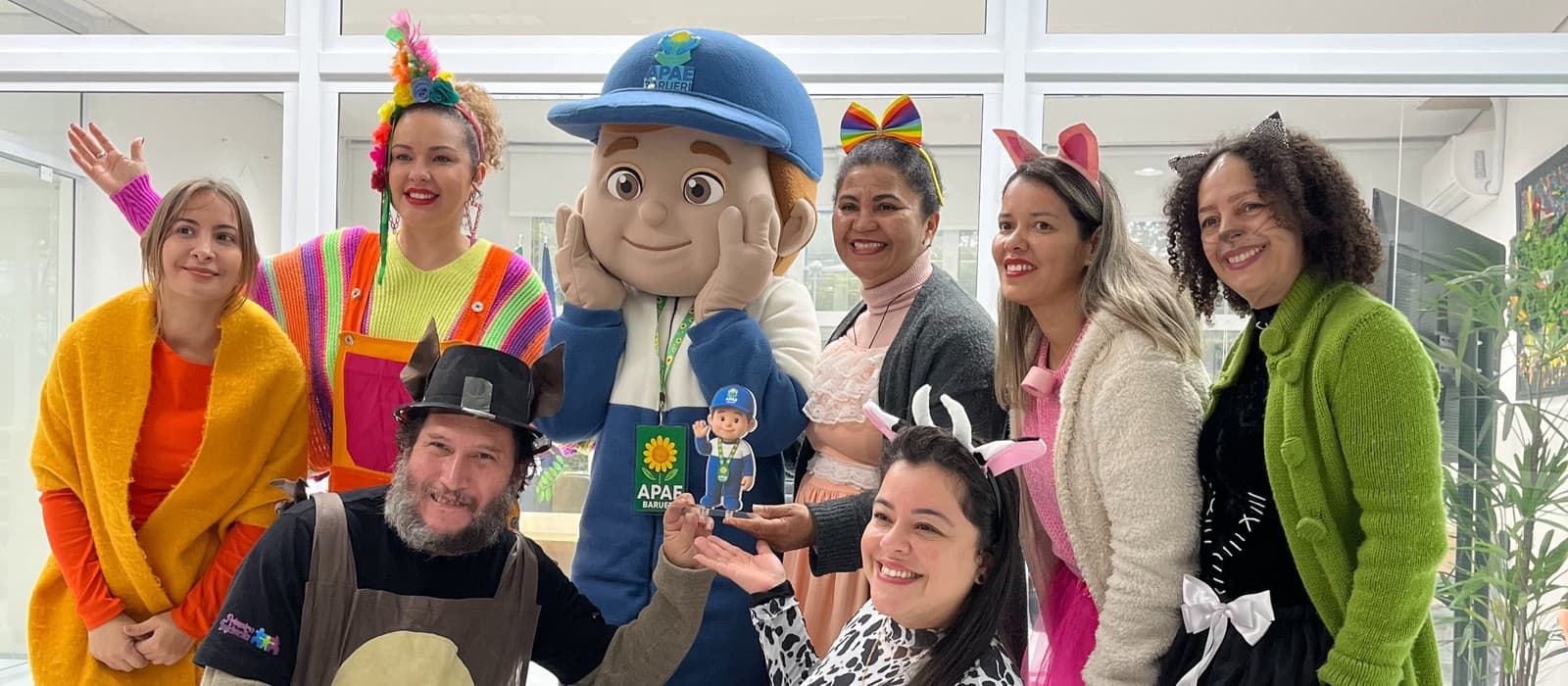 Oito adultos fantasiados ao redor do mascote APAEzito em tamanho real, que segura o Troféu APAEzito de Ouro — uma placa de acrílico com o personagem acenando —, num ambiente interno com divisórias de vidro.