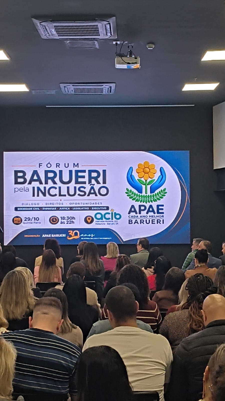 Auditório da ACIB lotado, visto do fundo da sala: a plateia, sentada de costas para a câmera, assiste ao telão que exibe a arte do Fórum Barueri pela Inclusão, com o tema Diálogo, Direitos, Oportunidades, a data 29/10, das 18h30 às 22h, e o símbolo da APAE Barueri, com mãos azuis envolvendo uma flor amarela.