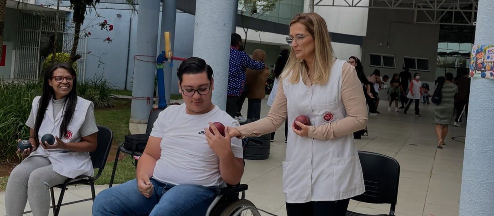 No pátio coberto da APAE Barueri, um jovem de óculos em cadeira de rodas segura uma bola vermelha de bocha, enquanto uma profissional de colete branco, de pé ao lado dele, segura outra bola; à esquerda, outra profissional sentada sorri com duas bolas azuis nas mãos.
