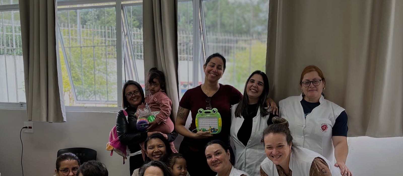 Profissionais da APAE Barueri, de avental branco com o símbolo da instituição, reunidas numa sala ampla com atendidos e adultos que os acompanham, todos sorrindo para a foto. À frente, uma mesa coberta de brinquedos embalados em papel celofane com laços: bonecas, bichos de pelúcia, carrinhos e um brinquedo de encaixar com peças coloridas. Ao fundo, janelas com cortinas bege.