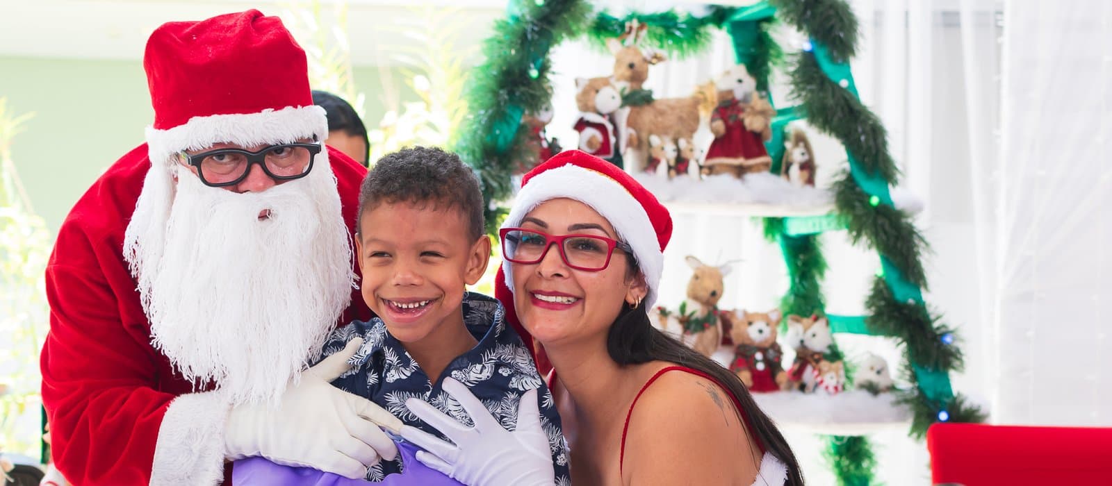 Na Festa de Natal da APAE Barueri de 2023, um menino sorridente, de camisa estampada, segura um presente embrulhado em papel lilás, abraçado por um Papai Noel de óculos e por uma mulher de gorro de Natal e óculos vermelhos; ao fundo, uma estante decorada com renas e bonecos natalinos.