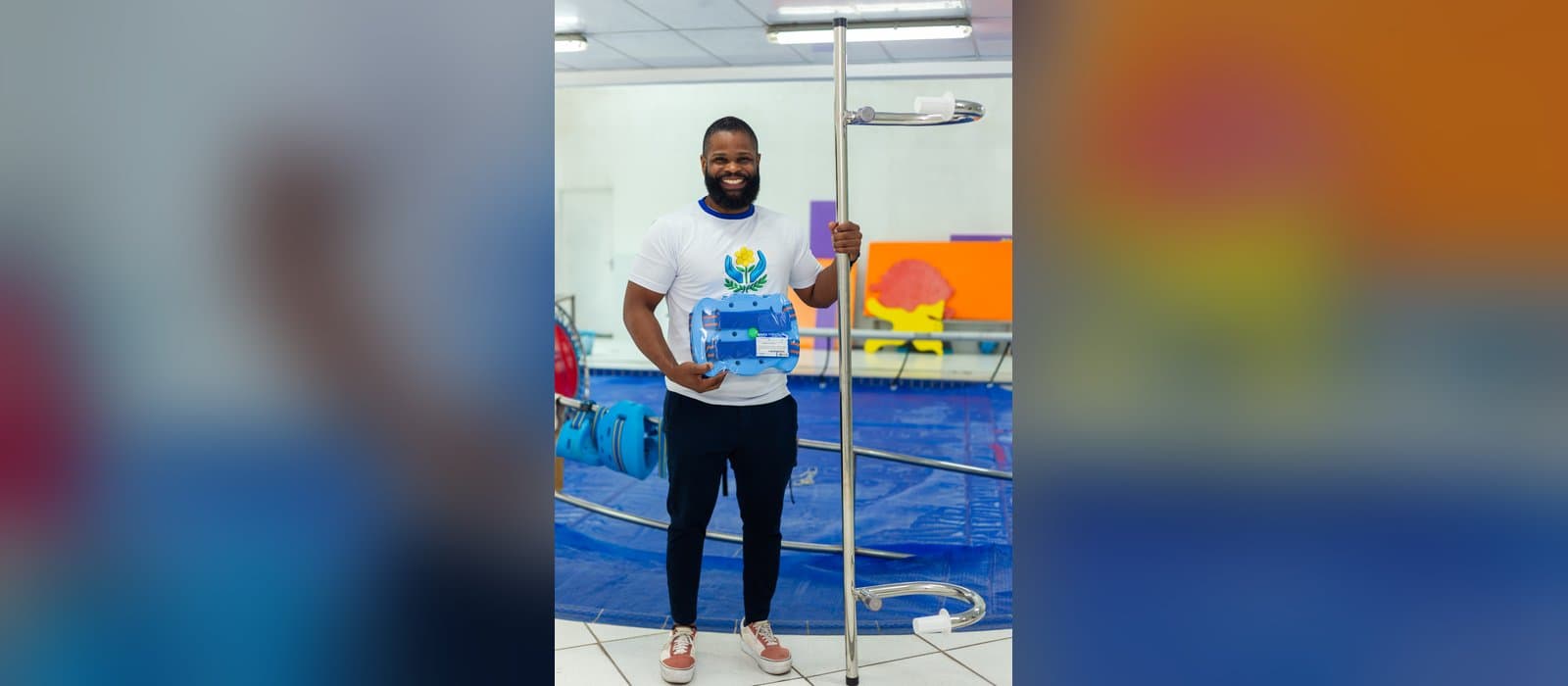 Profissional da APAE Barueri, de camiseta branca com o símbolo da instituição, sorri de pé à beira da piscina, segurando com uma mão um flutuador azul ainda na embalagem e com a outra uma barra de apoio de inox; atrás dele, a piscina coberta por uma lona azul, boias penduradas no corrimão e painéis coloridos na parede.